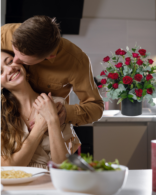 Romantic floral arrangement of red roses and fresh greenery in a sleek black vase, placed in a cozy dinner setting. A perfect gift for anniversaries, date nights, and heartfelt surprises.
