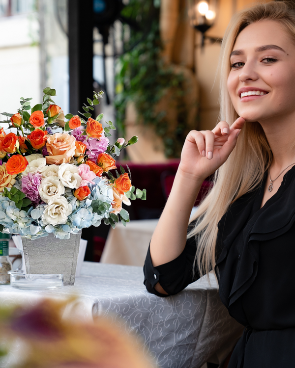 Color Splash floral arrangement featuring blue hydrangeas, orange spray roses, light peach roses, purple stock, and eucalyptus in a stylish wooden vase. A bright and cheerful gift for any occasion.